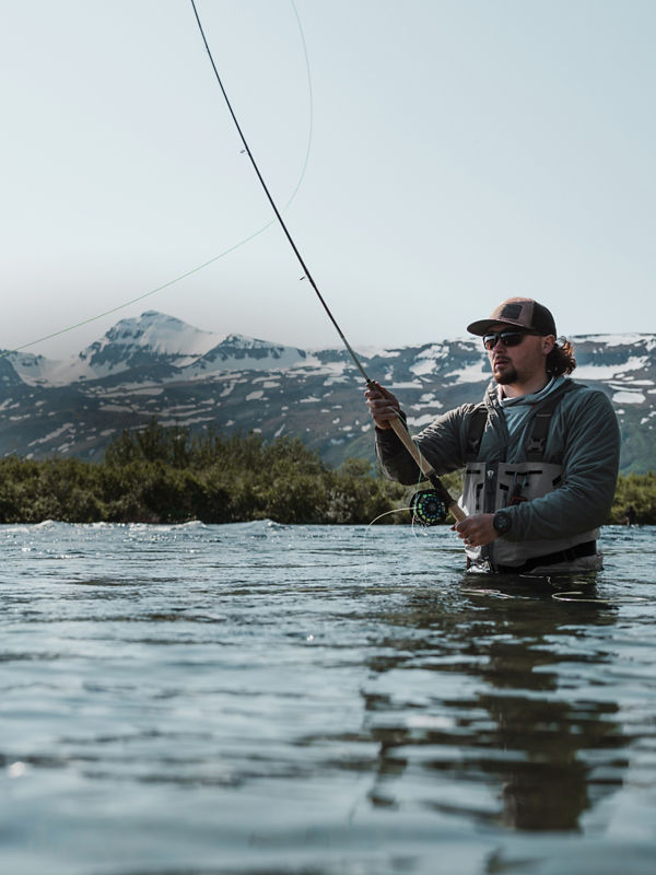 An angler in waders casts from waist-deep in a wide river.