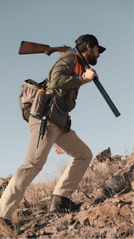 A man climbing a hill wearing his hunting gear and carrying a shotgun.