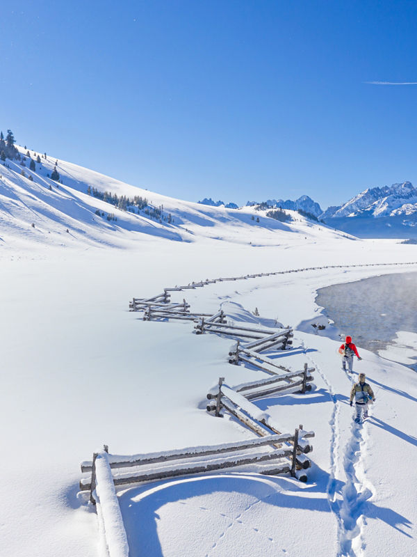 Two people forging a path through deep snow to an iced-over river for fly fishing.