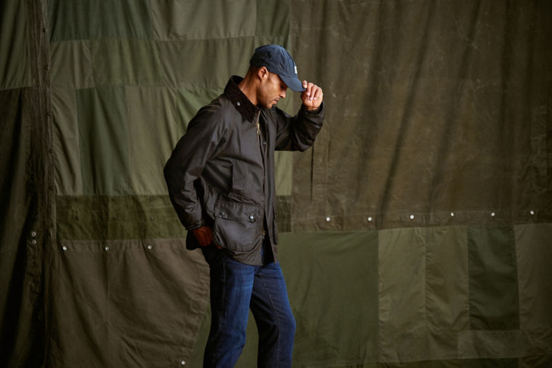 A man stands in front of a green backdrop wearing a Barbour waxed jacket and a ball cap.