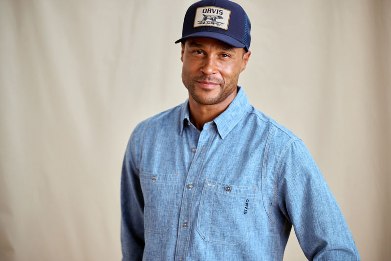 A model stands in front of a canvas drop cloth wearing a denim blue button-down shirt and ball cap.