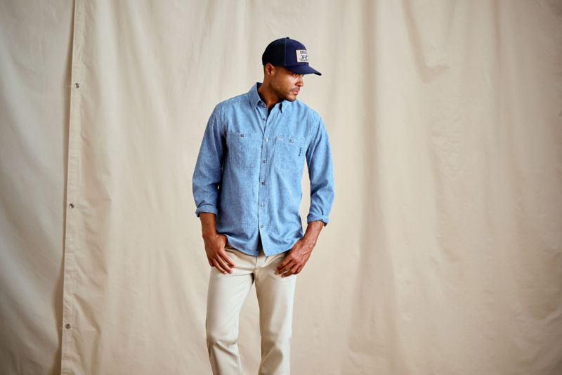 A model wears a blue tech chambray button-down shirt with a navy ball cap.