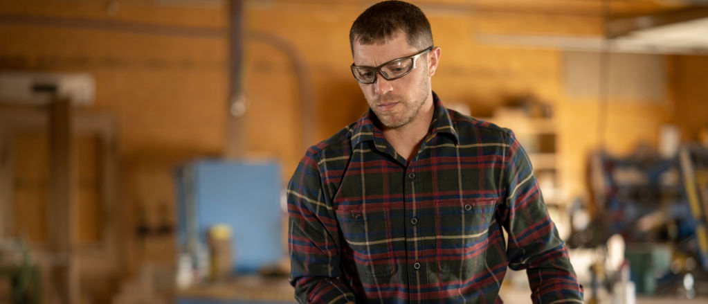 Man wearing a red flannel shirt working in a wood shop.
