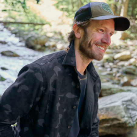 A smiling man in a black-and-grey camo jacket stands in front of a rocky stream.