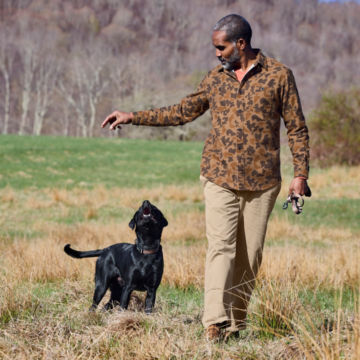A man wearing a camo shirt while walking with his black dog