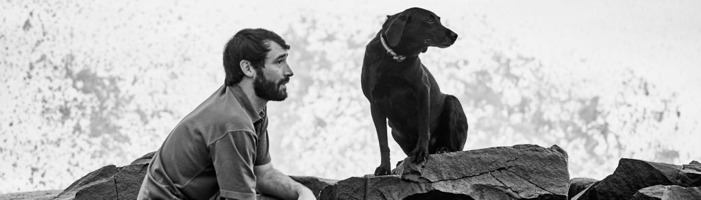 A black and white photo of a hiker resting on a pile of rocks with his black Labrador Retriever.