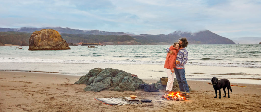 A man and a woman hugging on a beach in front of a small fire with their dog
