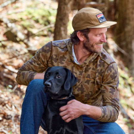 A man in '71 camo kneels in the woods next to his alert black Labrador