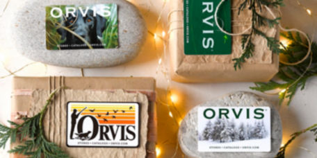 Orvis gift cards on a plaster background with wrapped gifts, river rocks, and fir boughs