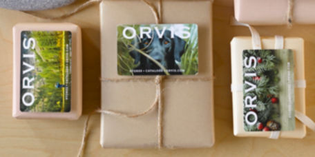 An assortment of small paper-wrapped gifts with Orvis Gift Cards on top and stones interspersed.