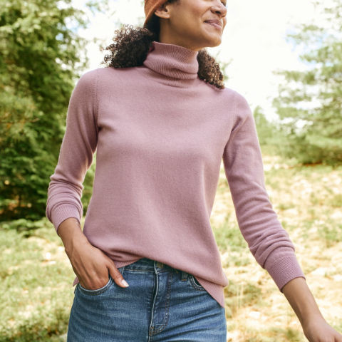 A woman in a pink cashmere turtleneck with jeans poses for the camera in the outdoors