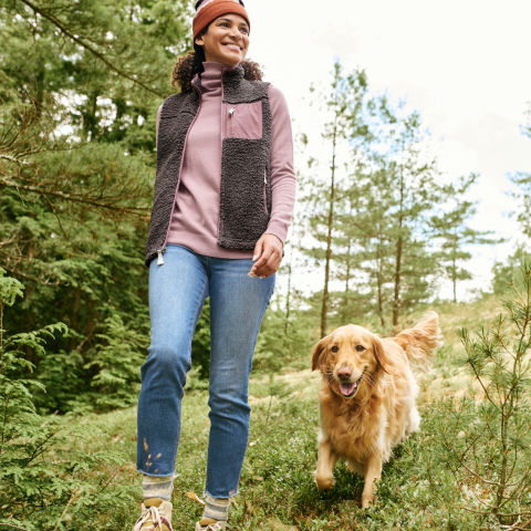A woman in the pink cashmere turtleneck walks through the woods with a golden retriever
