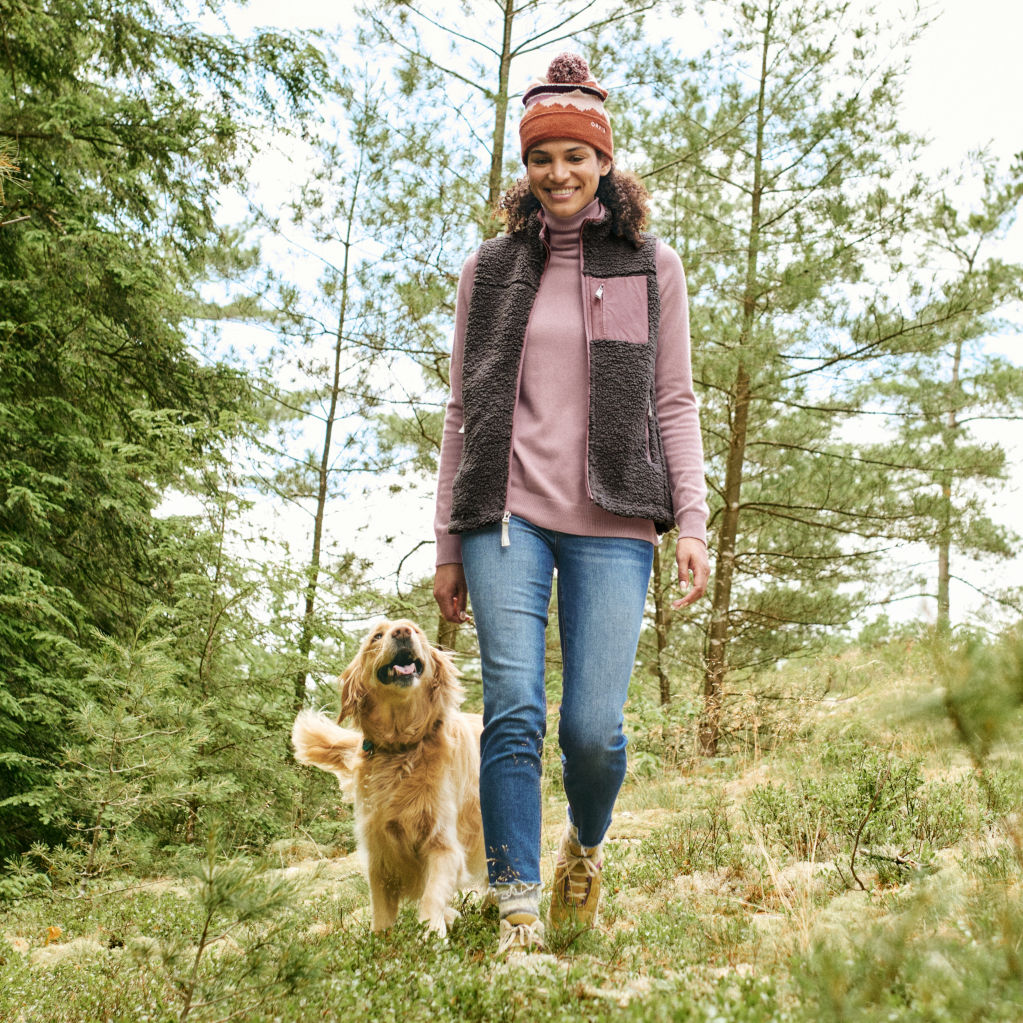 A woman in the pink cashmere turtleneck walks through the woods with a golden retriever