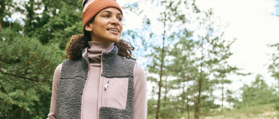 A woman in a gray fleece vest and pink cashmere turtleneck walks through a forest