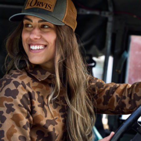 A woman wearing the '71 camo flannel shirt and a trucker cap, sitting behind the steering wheel of a vintage truck