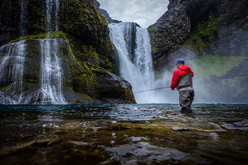 An angler in waders fly fishes in front of a waterfall in Iceland. 