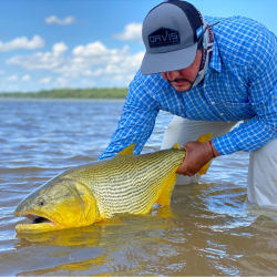 A man wearing a hat and blue checked shirt in the water holding a large yellow fish
