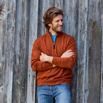 Man in Signal Orange Merino Wool Quarter-Zip Sweater 2.0 leans against a wooden wall.