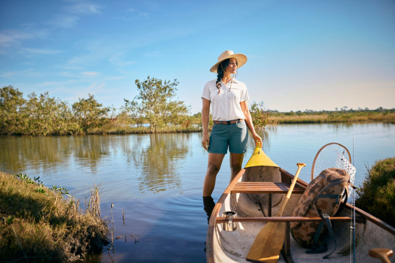 Woman in White Open-Air Caster Short-Sleeved Shirt wades in marsh next to rowboat.