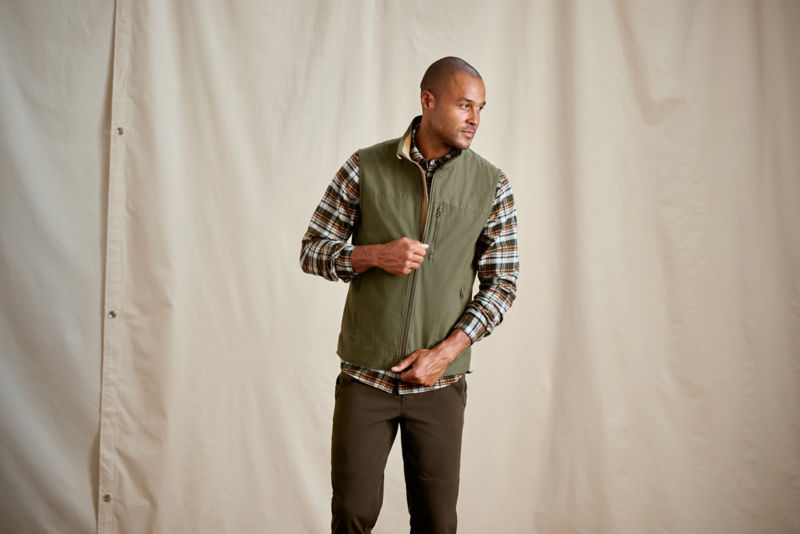 A model zipping an olive green vest over a plaid flannel shirt.