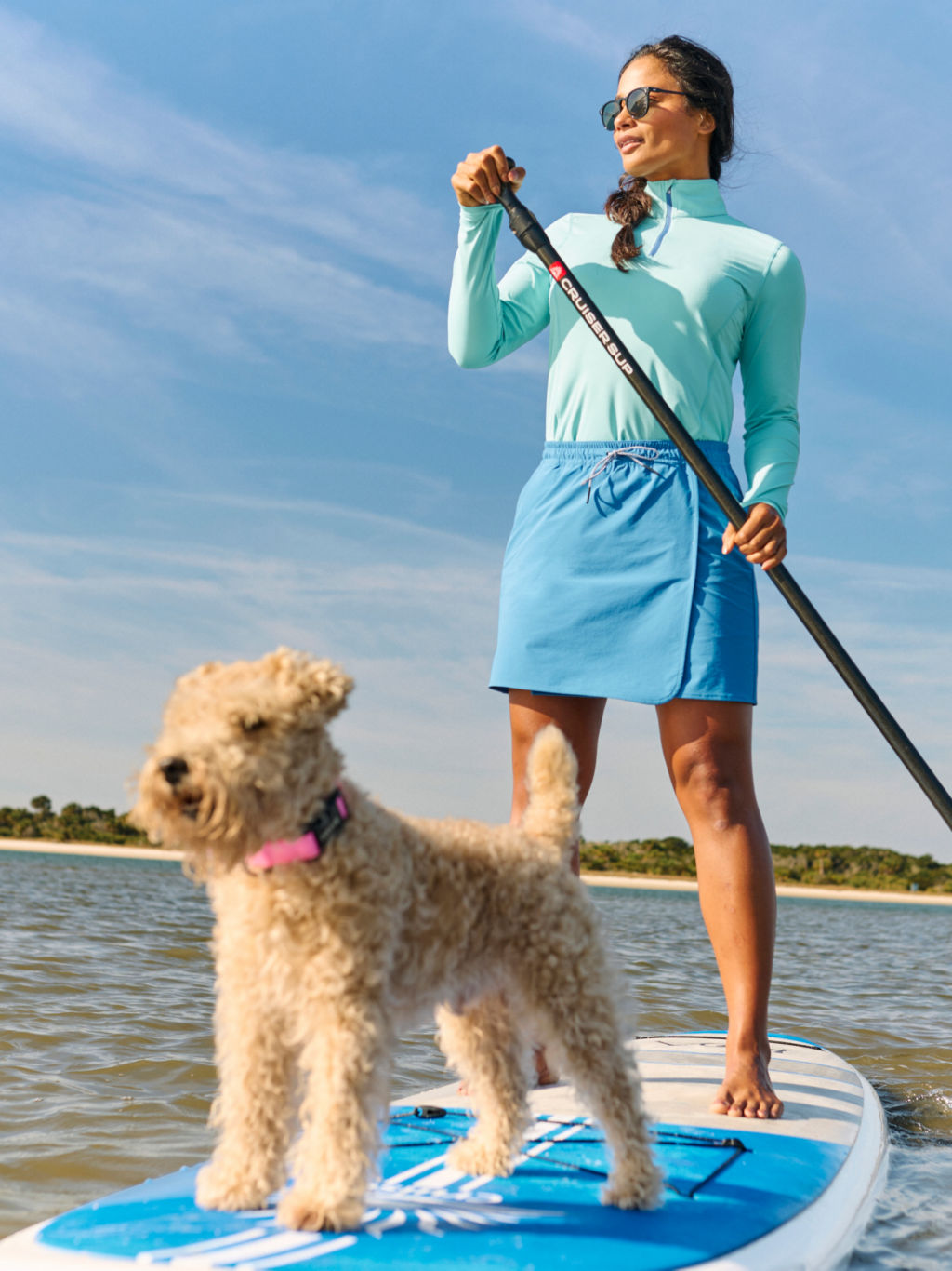 A woman wearing turquoise sun protection clothing stands on a paddle board with her fluffy-coated dog.