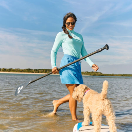 A woman wearing sun-protective clothing, holding a paddle board paddle, and walking along a beach with her dog.