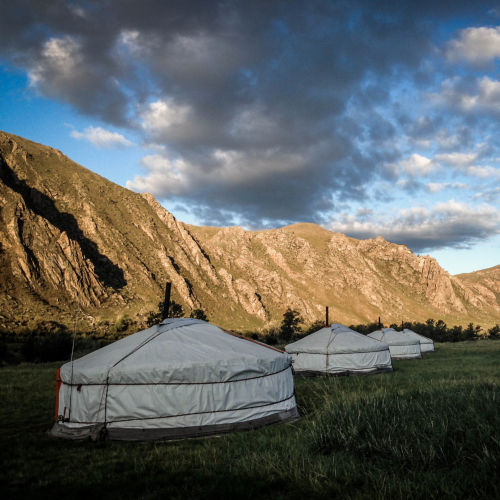 White yurts rest beneath angry clouds with rocky mountains in the background.