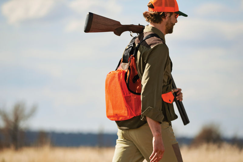 A hunter wearing a Waxed Cotton Strap Vest walks through high brush.