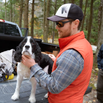 Man pets dog in bed of truck as angler group gathers in the woods.
