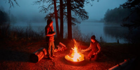 Two campers huddle around a blazing campfire next to a lake.