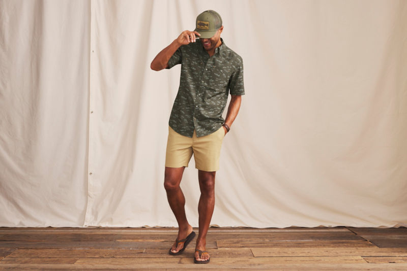 A smiling man adjusts his olive ball cap while wearing a patterned green short sleeve button-down shirt.
