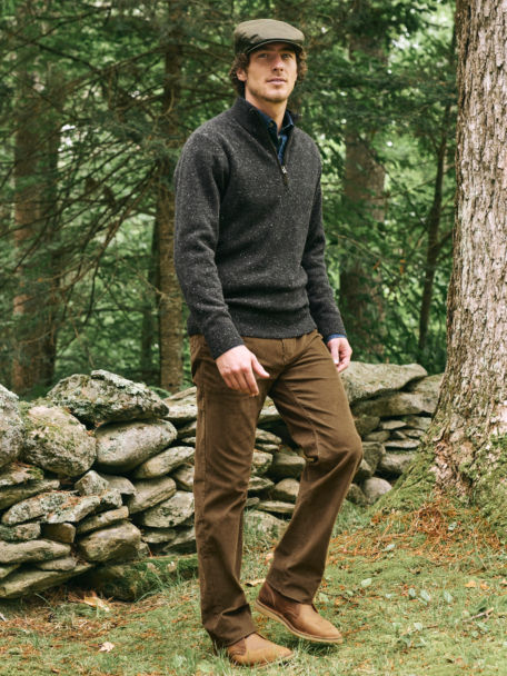 Man in Charcoal Merino Donegal Quarter Zip walks along a stone wall on the outskirts of a forest.