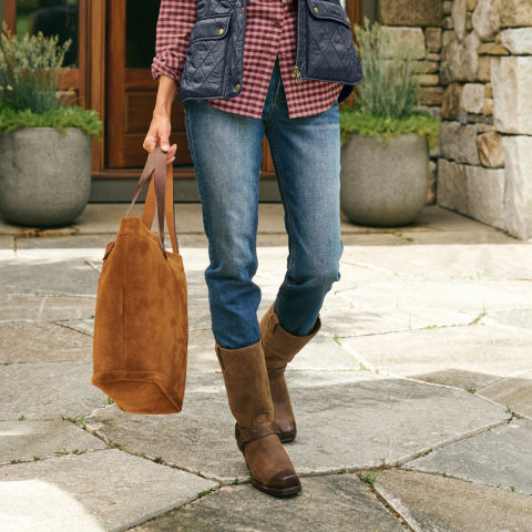 A woman in tall leather boots with a suede tote walks across a stone patio