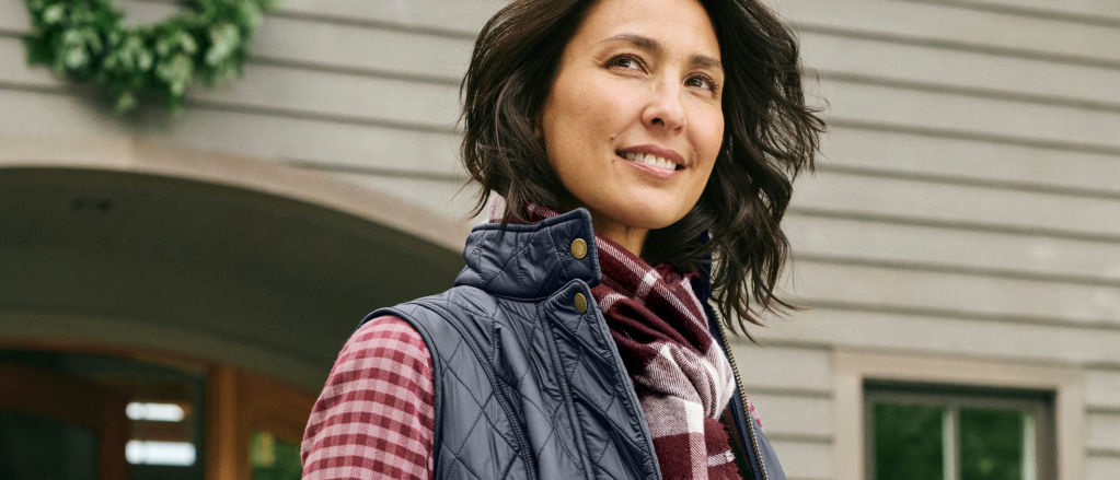 A woman in a navy quilted vest and red flannel shirt stands in front of a house with a wreath of greenery.