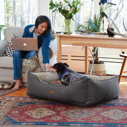 A woman leaning down to pet her dog who is laying in bolstered dog bed