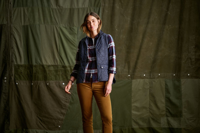 A model wearing a navy quilted vest over a plaid flannel shirt.