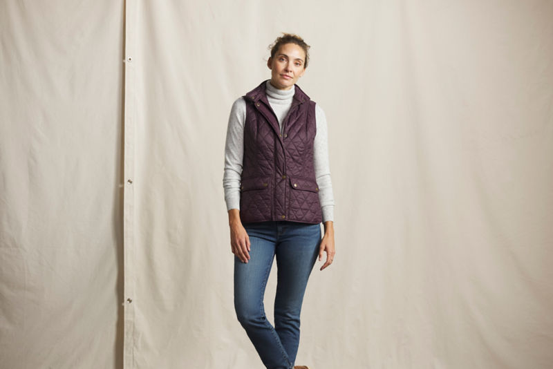 A model wearing a cranberry quilted vest over a white turtleneck and blue jeans.