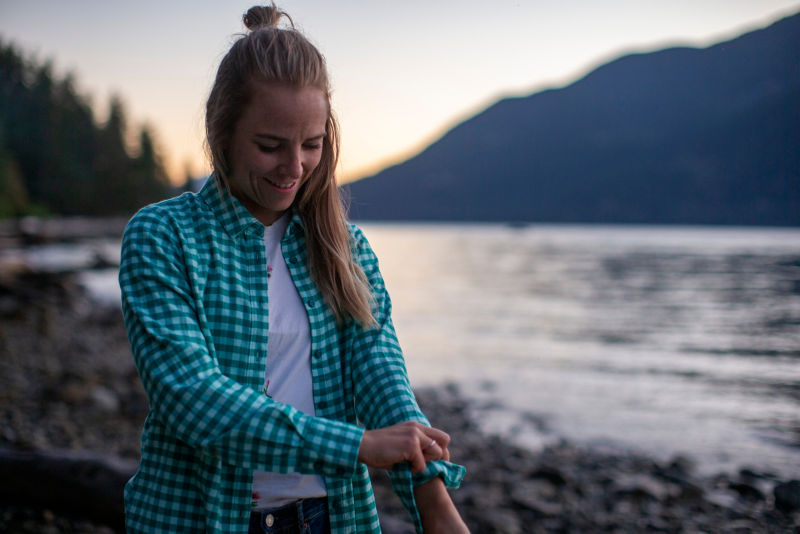 A hiker pauses on a lakeshore wearing a teal checked shirt.