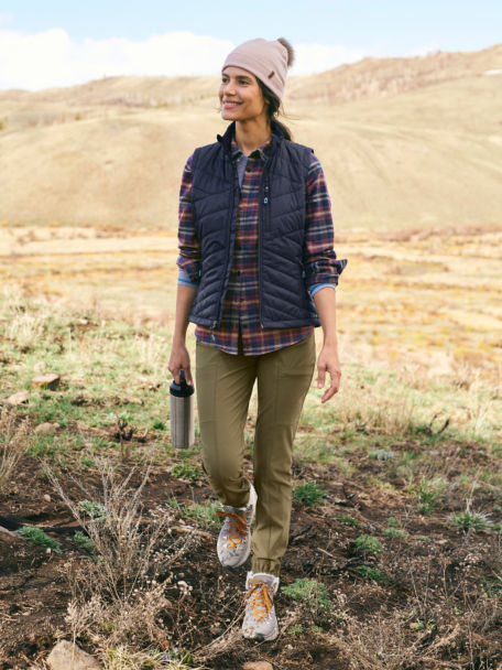 Woman in Navy Tech Flannel Shirt and Vest walks along a grassy knoll with her water bottle.