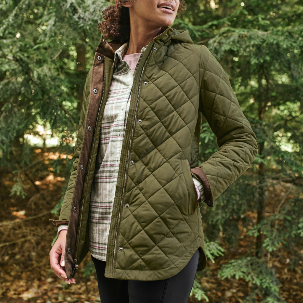 A woman stands in a forest wearing a green flannel shirt and an olive quilted coat with hood