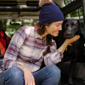 Woman in Lodge Flannel Plaid Shirt sits in the back of her land rover and pets her dog.