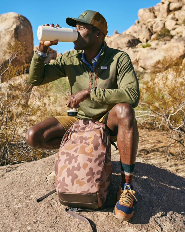 A man squatting down in a desert drinking from a white thermos.