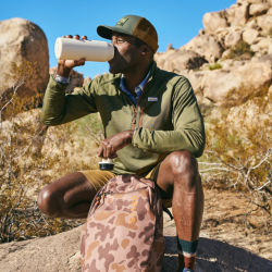 A man squatting down in a desert drinking from a white thermos.