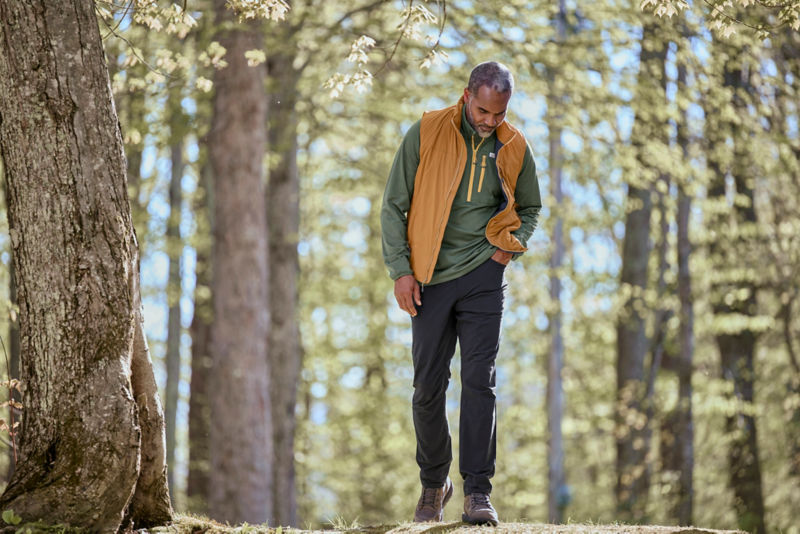 A model walks a forest path wearing a gold vest over a green long-sleeve t-shirt.