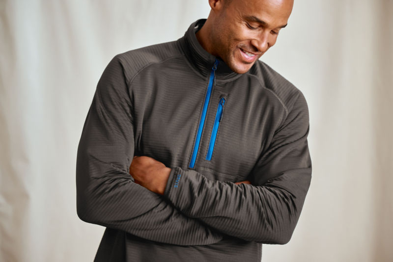 A model stands in front of a canvas backdrop wearing an olive sweatshirt with a bright blue zipper up the front.