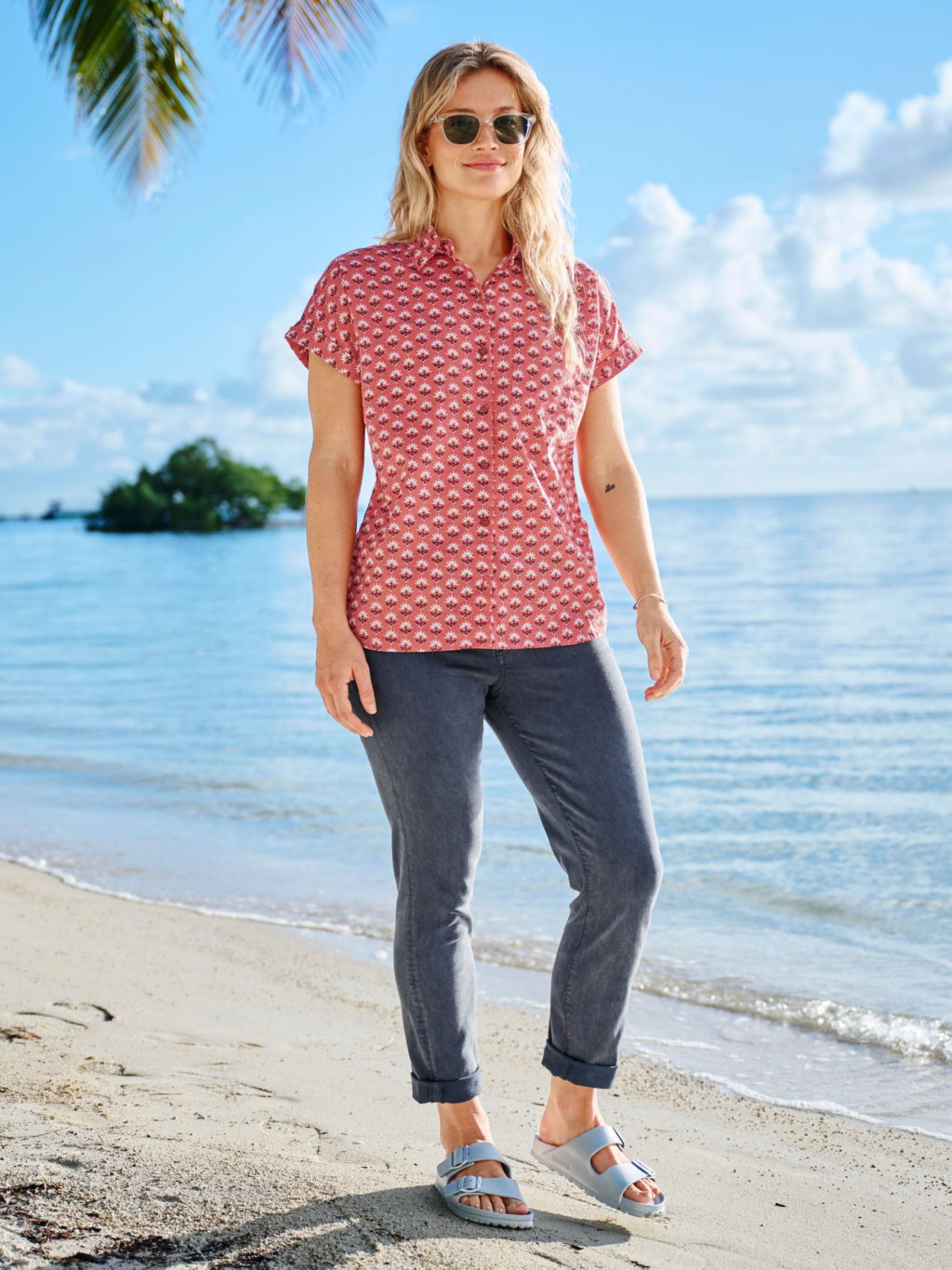 A blonde woman wearing sunglasses, a red patterned short sleeved button down shirt and deep navy pants.