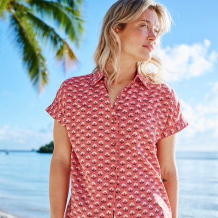 A woman wearing a red printed shirt-sleeved button down shirt, standing on a tropical beach