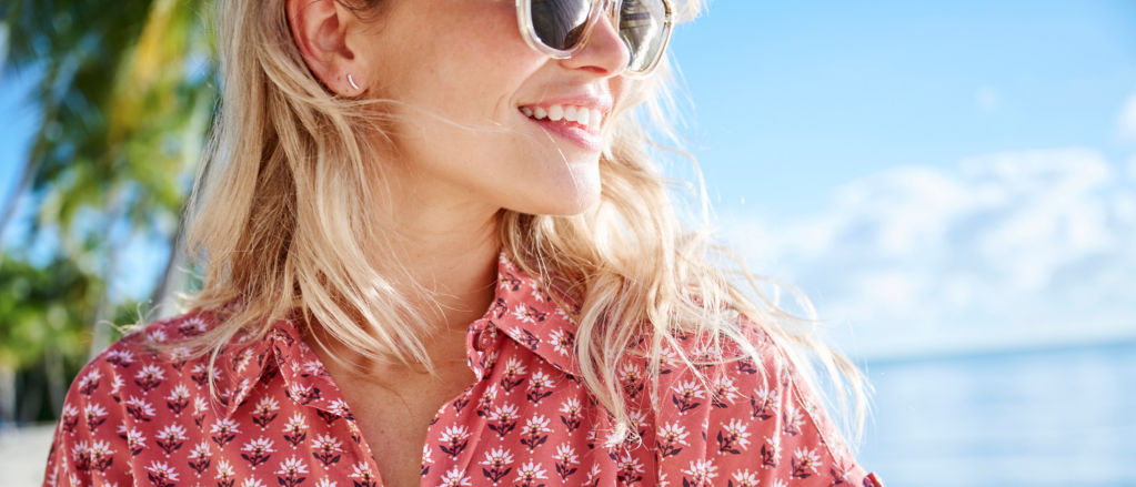 A close-up of a blonde woman's profile wearing sunglasses on a tropical beach.