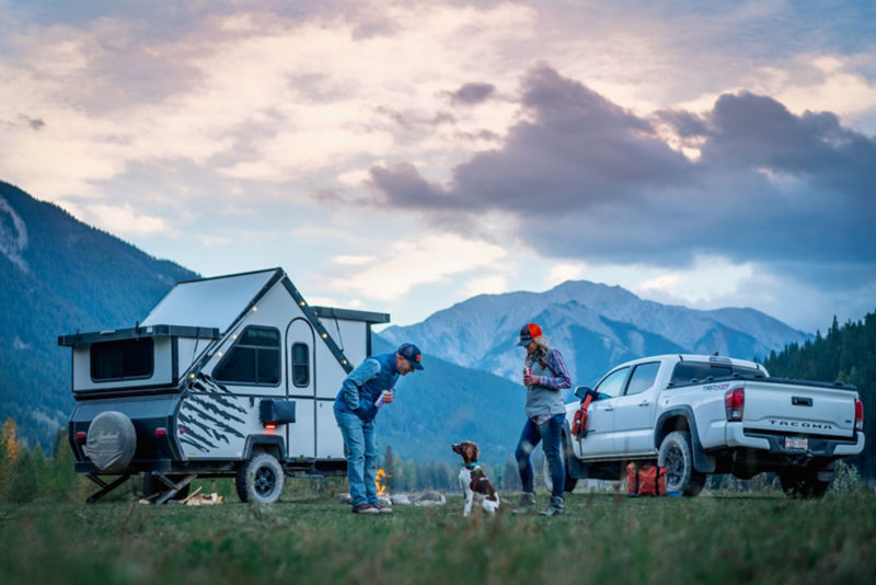 A man, woman, and their dog in front of their white truck and white camper in the great outdoors