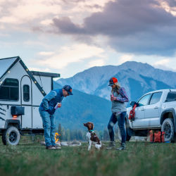 A man, woman, and their dog in front of their white truck and white camper in the great outdoors